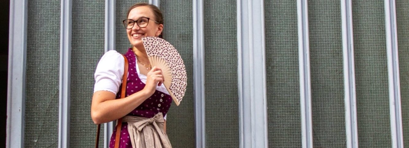 Aufnahme einer Person mit Trachtenfächer und Bordeaux-farbenem Dirndl vor einer Glaswand